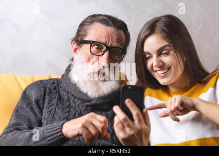 Surpris dans les verres hauts père barbu assommé l'erreur ou l'erreur message lors de l'utilisation de smartphone, il s'assis sur le canapé jaune à la lumière salon avec sa fille brune aux cheveux long adultes, elle sourit et explique à son père comment il fonctionne, la technologie moderne, communication concept Banque D'Images