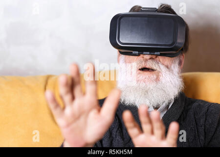 Homme barbu senior surpris en pull gris touché quelque chose avec ses mains lors du test de l'appareil VR, il s'assis sur le canapé jaune dans la pièce vivante de lumière, avenir technologie concept Banque D'Images