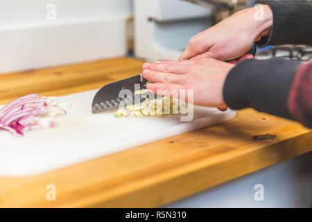 Caucase woman manches longues coupe les morceaux de l'ail sur une planche à découper en plastique. Oignons rouges tranchés à proximité. Banque D'Images