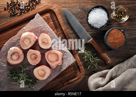 Les os de veau brut blanc sur papier de cuisson et planche à découper en bois. Décorées de fines herbes, d'épices, couteau du chef et une serviette. Vue de dessus. Banque D'Images
