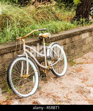 Un vieux beach bike garé contre un mur de pierre a un pneu à plat, reposant sur le sable. Banque D'Images
