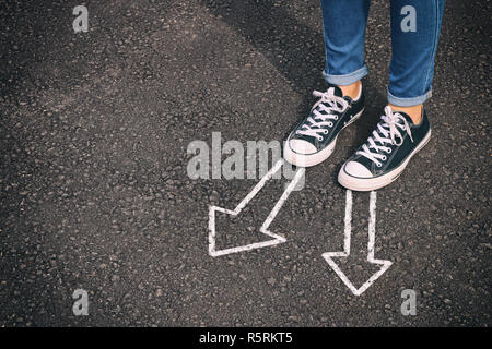 Haut voir l'image de la personne en jeans et chaussures rétro debout au-dessus de la route d'asphalte peint avec des flèches indiquant des directions différentes Banque D'Images