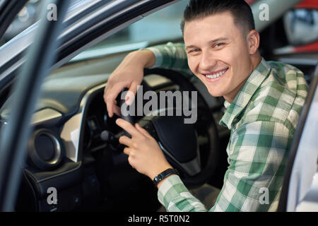 Portrait de jeune homme assis sur la nouvelle voiture sur le siège conducteur. Guy attrayant en chemise à carreaux à la caméra et au sourire. Heureux propriétaire de véhicule maintenant ses mains sur le volant. Banque D'Images