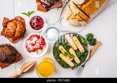 Petit déjeuner servi avec des muffins, des saucisses grillées, jus de fruits, pain frais et parfait sur une table en bois blanc, vue du dessus. Banque D'Images