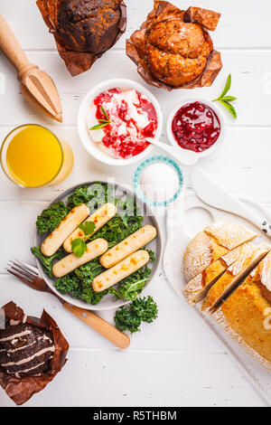 Petit déjeuner servi avec des muffins, des saucisses grillées, jus de fruits, pain frais et parfait sur une table en bois blanc, vue du dessus. Banque D'Images