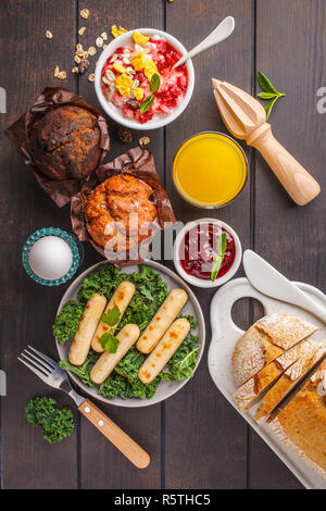 Petit déjeuner servi avec des muffins, des saucisses grillées, jus de fruits, pain frais et parfait sur une table en bois sombre, vue du dessus. Banque D'Images