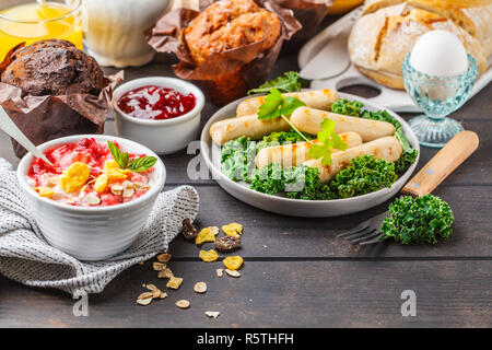 Petit déjeuner servi avec des muffins, des saucisses grillées, jus de fruits, pain frais et parfait sur une table en bois sombre. Banque D'Images