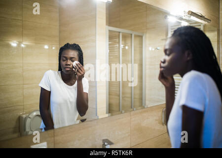 Gardant sa peau douce et lisse. Belle jeune femme africaine face nette des produits de beauté et de sourire tout en se tenant contre un miroir dans la salle de bains Banque D'Images