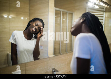 Gardant sa peau douce et lisse. Belle jeune femme africaine face nette des produits de beauté et de sourire tout en se tenant contre un miroir dans la salle de bains Banque D'Images