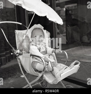 Années 1960, historique, d'une journée ensoleillée, et un bébé portant un bonnet à l'extérieur de certains magasins dans un petit encadré de métal poussette avec un parasol parasol. Banque D'Images