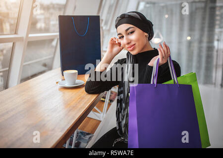 Beau jeune arabian model posing sur appareil photo. Elle s'assoit à la table près de l'intérieur de la fenêtre dans le café. Jeune femme tenir les sacs à main avec Banque D'Images