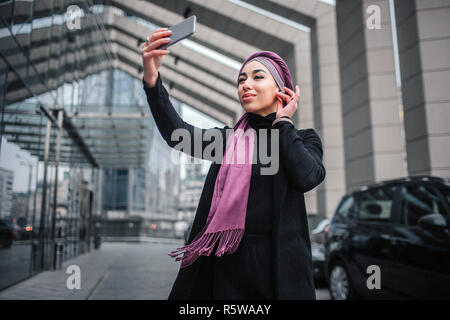 Gai et attractive young woman in hijab selfies prendre. Modèle d'Oman se trouve à l'extérieur. Elle est titulaire d'main près à la tête Banque D'Images