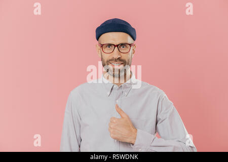 Satisfait l'homme a beau look attrayant, soulève le pouce, d'accord avec quelque chose, sourit joyeusement, porte un chapeau et chemise, plus isolé sur fond rose. Banque D'Images