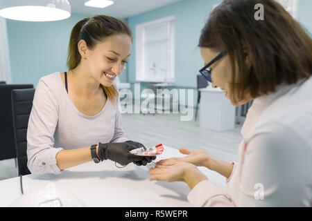 Jeune fille esthéticienne manucure parle à une cliente, manucure, ne choisit la couleur de vernis à ongles, communique et sourit. Banque D'Images