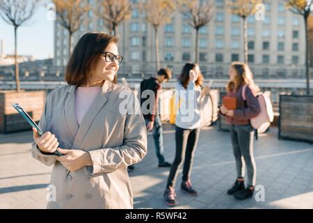 Portrait of mature female teacher dans les verres avec des presse-papiers, piscine avec un groupe d'Adolescents Étudiants, heure d'or. Banque D'Images