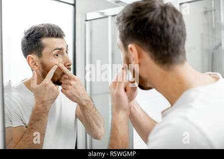 Homme en blanc T-shirt comprimant l'acné sur son visage à la recherche d'un miroir dans la salle de bains Banque D'Images