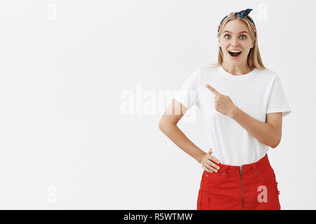 Tourné à l'intérieur d'impressionné et intrigué heureuse femme slim attrayant avec des cheveux blonds en jupe rouge et serre-tête vers le coin en haut à gauche et souriant Banque D'Images