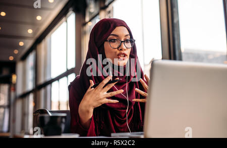 femme d'affaires dans le hijab ayant un chat vidéo sur ordinateur portable tout en étant assis au café. Femme assise au café et faisant un appel vidéo en utilisant des écouteurs et la Banque D'Images
