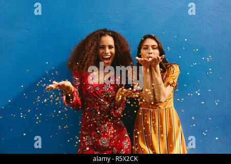 Female friends blowing off magic glitter. Les femmes excitées s'amusant contre mur bleu. Banque D'Images