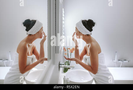 Serviette femme enveloppée dans l'application de la crème sur le visage, debout devant un miroir. Asian woman grooming dans salle de bains. Banque D'Images