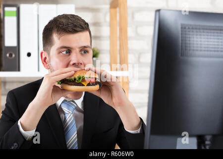 Business Man Eating Burger while looking at Computer Banque D'Images