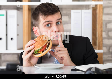 Faim Young Businessman Holding Burger avec le doigt sur les lèvres Banque D'Images