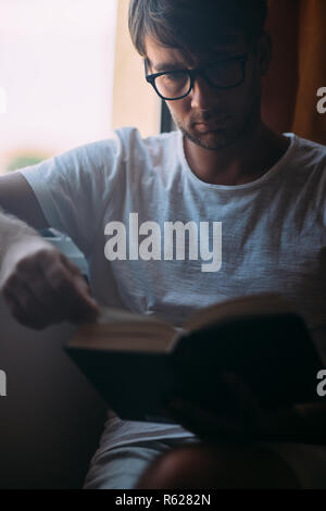Jeune homme séduisant en lunettes lecture livre assis près de la fenêtre. week-end concept Banque D'Images