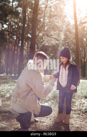 Jeune mère aide sa petite fille à porter curly coat pendant qu'ils marche dans les bois. La saison froide, clair soleil est visible à travers les arbres en forêt Banque D'Images