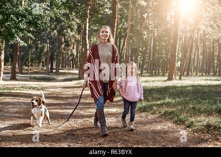 Mère et fille avec chien randonnées en forêt de pins. Femme porte un élégant manteau en tricot, fille est en pull rose Banque D'Images