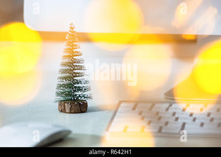 Un petit sapin se dresse sur un bureau blanc, près de l'écran d'un ordinateur, clavier et souris. Concept, créer une ambiance de Noël sur le lieu de travail. Banque D'Images
