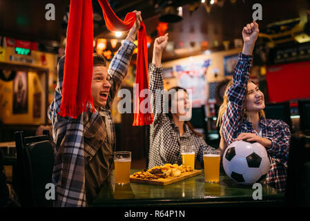 Fans de football avec écharpe regarder match et lever la main jusqu'au bar des sports. La diffusion Tv, jeunes amis loisirs en pub, équipe favorite gagne Banque D'Images