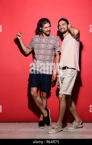 Portrait d'une deux jeunes frères jumeaux joyeux sur fond rouge isolé, écouter de la musique avec des écouteurs Banque D'Images