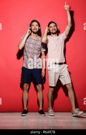 Portrait d'une deux jeunes frères jumeaux joyeux sur fond rouge isolé, écouter de la musique avec des écouteurs, la danse Banque D'Images