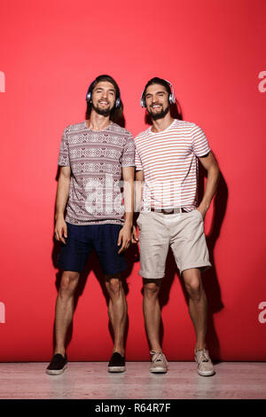 Portrait d'une deux jeunes frères jumeaux joyeux sur fond rouge isolé, écouter de la musique avec des écouteurs, looking at camera Banque D'Images