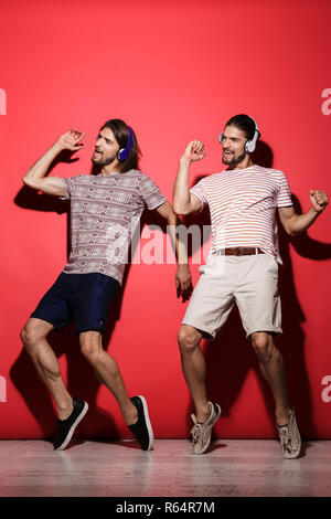 Portrait d'une deux jeunes frères jumeaux joyeux sur fond rouge isolé, écouter de la musique avec des écouteurs, danser, s'amuser Banque D'Images