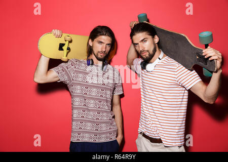 Portrait de deux jeunes frères jumeaux attrayants sur fond rouge isolé, écouter de la musique avec des écouteurs, holding skateboards Banque D'Images