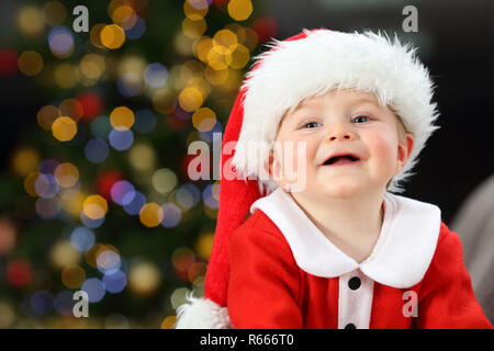 Kid looking at camera wearing santa claus déguisement sur un canapé à Noël avec un arbre dans l'arrière-plan Banque D'Images