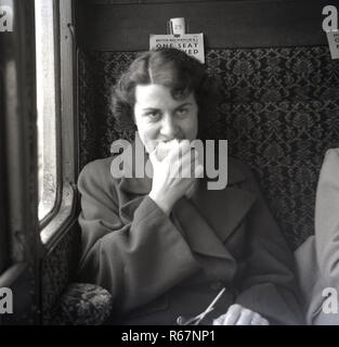 Années 1950, historiques, une jeune femme assise dans son manteau dans un compartiment de train à côté de la fenêtre de manger une pomme. Banque D'Images