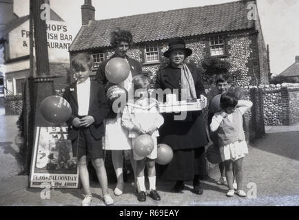 Vers 1930s, historique, à l'extérieur d'un village anglais, une journée « drapeau et bordeaux »... deux mères avec leurs jeunes enfants tenant des ballons pour la collecte de fonds de la « Shipwrecked Mariners Society ». Depuis 1839, l'organisme de bienfaisance a fourni un soutien aux marins et aux pêcheurs marchands à la retraite et, bien sûr, à ceux qui sont touchés par les naufrages. Affiche pour l'événement sur le terrain à côté d'un garçon, notez l'orthographe du ballon comme 'bordeaux'. Banque D'Images