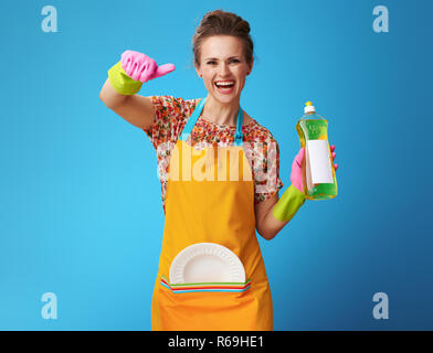 Happy housewife en orange tablier avec plaque lavé et détergents showing Thumbs up isolé sur le bleu ! Femme au foyer montre favorite Banque D'Images