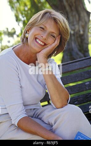 Portrait, Parkszene, blonde Frau, Mitte, 50 bekleidet mit heller und flexible Top, sitzt entspannt mit einem Buch in der Hand auf einer Parkbank unter einem Baum