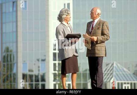 Seniorenpaar, gepflegter Herr mit Anzug und roter Krawatte senneur mit Gattin grauen im bekleidet Kostuem Glasfassade vor einer mit Dokumenten und Tageszeitung Banque D'Images
