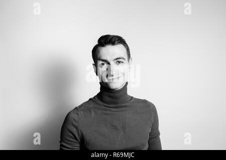 Close up portrait noir et blanc d'un jeune homme portant un cavalier col roulé, souriant à la caméra, contre un arrière-plan studio Banque D'Images