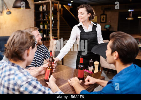 Happy smiling jolie fille en tablier noir debout à table avec les invités et les bouteilles de bière à donner en attendant leur table dans un bar de nuit Banque D'Images