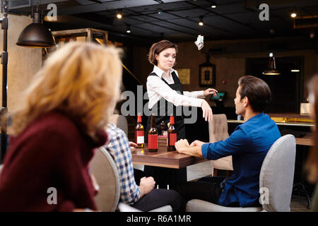 Mécontents agressifs attrayant jeune serveuse en tablier noir debout à table et en jetant de l'argent dans votre visage tout en exprimant sa négligence à Dar Banque D'Images