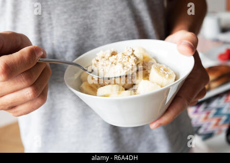 Libre d'un jeune homme portant un T-shirt gris occasionnels ayant un peu de porridge à la banane d'un bol en céramique blanche, debout à côté d'une table définie pour brea Banque D'Images