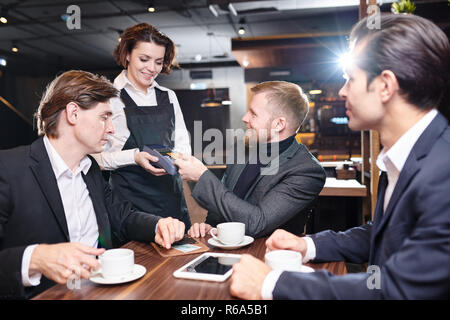 Jeune serveuse attrayant positive en tablier noir debout à la table des invités et donner à un terminal de paiement sans contact d'affaires qui désirent payer en c Banque D'Images