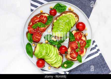 Du pain et des tomates à l'avocat des toasts avec du hoummos, sésame et basilic dans une plaque avec serviette sur fond de marbre blanc vegan aliments sains.. haut Banque D'Images