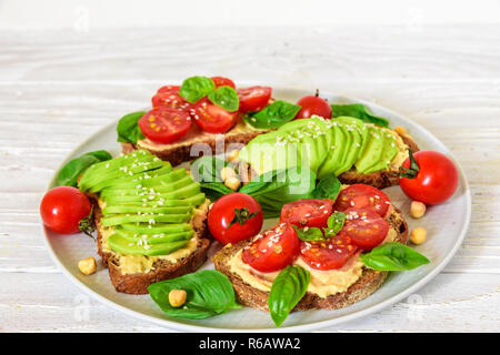 Du pain et des tomates à l'avocat des toasts avec du hoummos, sésame et basilic dans une plaque sur une table en bois blanc, en bonne santé. La nourriture végane. close up Banque D'Images