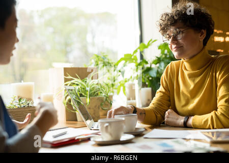 Confiant de contenu attrayant dame en col roulé jaune assis à table et de boire du café avec son associée dans le café moderne Banque D'Images